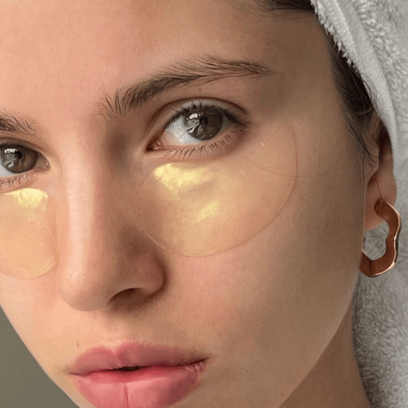 Woman applying gold eye patches with a towel on her head.